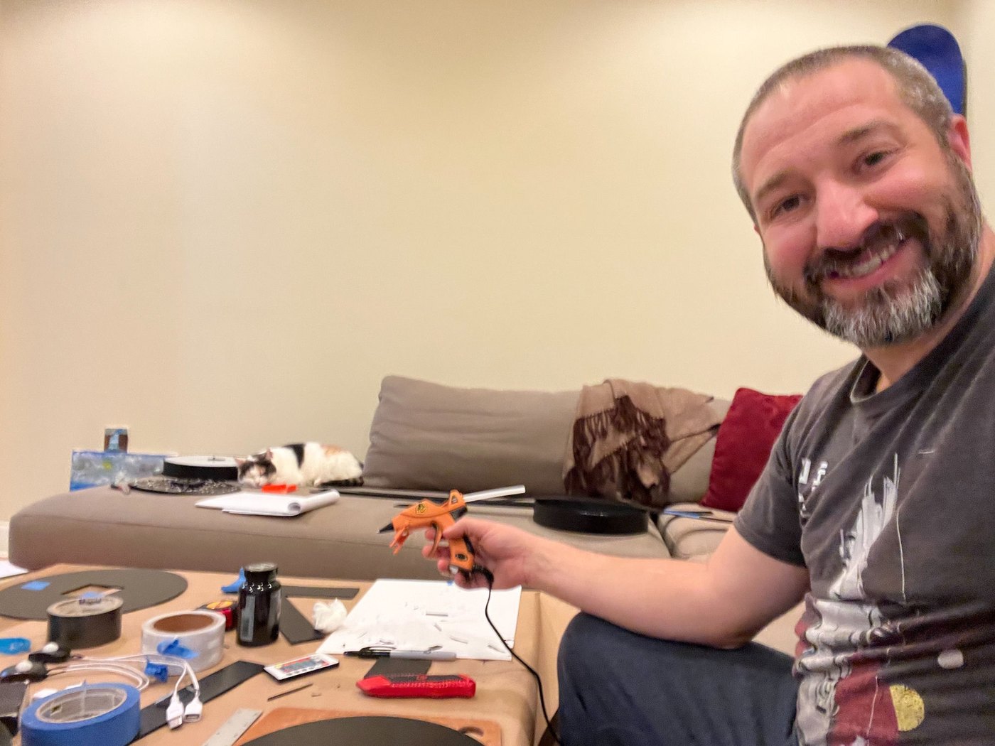 Andrew hand-making a Vylümi at his workbench, glue gun in hand, with his cat napping nearby